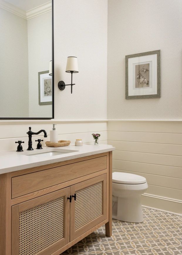 A refined powder room with a freestanding timber vanity featuring cane-panelled cabinet doors and a white countertop, paired with a dark traditional-style faucet and undermount sink. The room has cream horizontal wall panelling, patterned tile flooring, a large mirror, framed botanical artwork, and a toilet. A black wall sconce provides warm accent lighting.