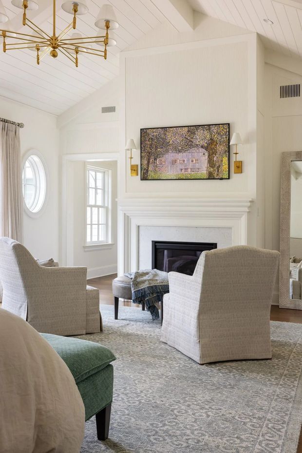 A bright, refined living room with white painted panelled walls and a vaulted, beadboard-style ceiling. The room is arranged around a white classical fireplace with a mounted television above it, upholstered armchairs, an ottoman, a patterned area rug, and a green upholstered sofa edge in the foreground. Tall windows and an oval window bring in natural light, while a brass multi-arm chandelier, brass wall lights, and a large decorative mirror add warmth and detail.