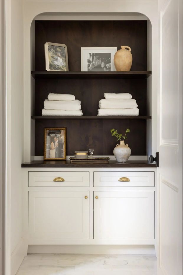 A built-in hallway storage and display cabinet with a dark wood recessed shelving niche, white painted cabinetry, brass hardware, folded towels, framed photographs, ceramic vessels, and a pale stone or marble floor. The cabinetry is framed by white paneled walls and a partially visible paneled door.