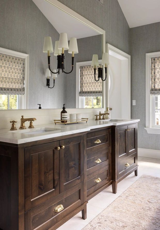 A refined bathroom featuring a large dark timber double vanity with paneled cabinetry, brass hardware and twin traditional faucets beneath a white stone benchtop. A large framed mirror reflects a black-and-brass multi-arm wall light, while grey patterned wallpaper, white trim, roman blinds and a pale rug create a classic, elegant interior.