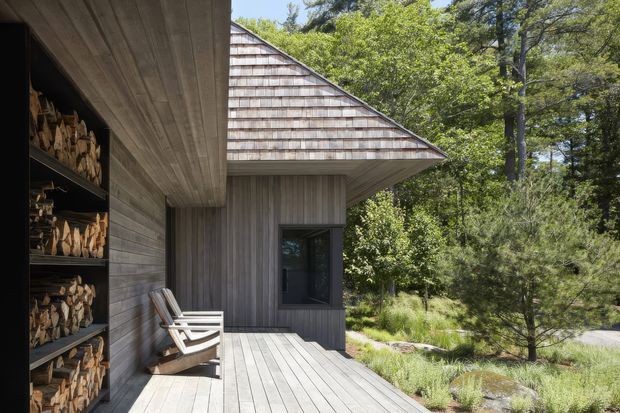 Exterior view of a small timber-clad structure in a wooded landscape. A broad timber deck runs along the facade, with a built-in open firewood storage wall and two outdoor lounge chairs beneath a deep roof overhang. The building has a simple gabled roof with shingle cladding on the gable end, vertical timber wall cladding, and a dark-framed window.