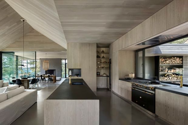 A contemporary open-plan kitchen viewed from the main cooking area, featuring a long dark island with integrated cooktop, pale timber cabinetry extending across the walls and ceiling, dark stone work surfaces, a large range cooker, open shelving, and a wide serving window. The kitchen opens directly into a dining and living area with extensive glazing and views to an outdoor wood storage feature.