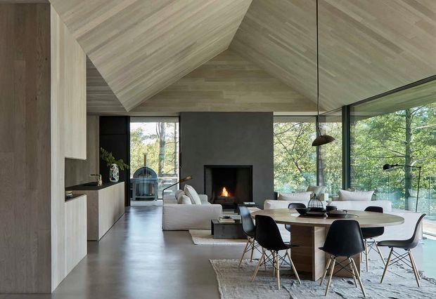 Open-plan contemporary living and dining area with a dramatic timber-lined vaulted ceiling, dark feature fireplace wall, extensive black-framed glazing, built-in kitchen cabinetry, a round dining table, and a light upholstered lounge. The room has a calm, natural connection to the surrounding trees and a refined neutral palette.
