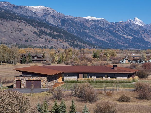 A low, single-storey contemporary house set in a rural mountain landscape. The home has a broad, low-pitched dark reddish-brown roof, extensive glazing, and a stone-clad garage wing and facade. Dry native landscaping surrounds the property, with forested foothills and snow-capped mountains forming the backdrop.
