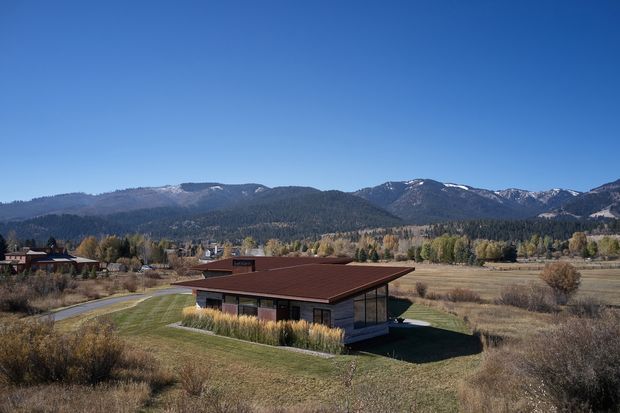Aerial exterior view of a low, contemporary single-storey home set in an expansive rural valley. The house has a broad shallow-pitched dark roof, timber-toned cladding, extensive glazing, and a simple rectangular form. Dry grassland, trees, a driveway, and forested mountains with light snow are visible in the surrounding landscape.