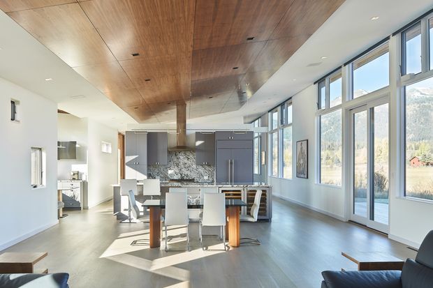 A bright, open-plan contemporary kitchen and dining area with a large island, white bar stools, dining table and chairs, extensive floor-to-ceiling windows, and a glazed door opening toward a scenic outdoor setting. The space features a warm timber-lined ceiling, pale flooring, white walls, grey kitchen cabinetry, and a patterned stone backsplash.