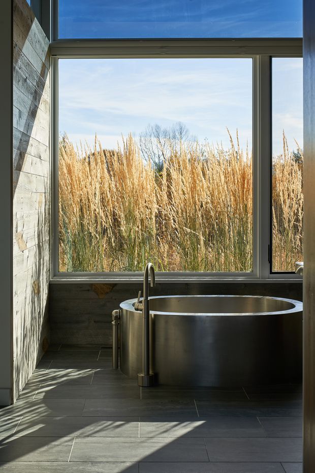 A serene bathroom with a freestanding metallic bathtub positioned beside large floor-to-ceiling windows. The room features dark grey tiled flooring, horizontal weathered timber wall cladding, dark-framed glazing, and a sculptural exposed bathtub faucet. Tall golden grasses and trees are visible outside, with strong sunlight casting long shadows across the floor.