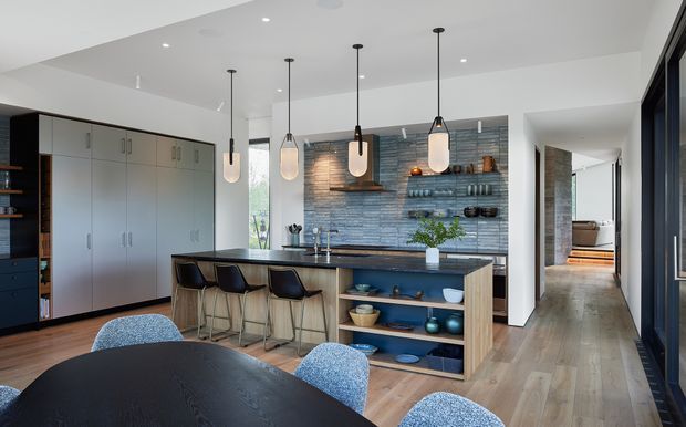 Bright contemporary open-plan kitchen and dining area with a large island, four pendant lights, extensive grey built-in cabinetry, and a textured blue-grey tiled feature backsplash with open shelving. A dark round dining table occupies the foreground, while a hallway and additional living space extend beyond the kitchen.