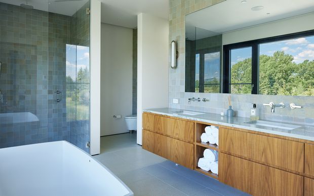 Spacious contemporary bathroom with a freestanding white bathtub in the foreground, a glass-enclosed tiled shower to the left, and a long double vanity with warm timber cabinetry and a pale stone benchtop. Large black-framed windows and a wide mirror bring in abundant natural light and views of greenery. The room has a calm, clean, refined atmosphere with muted blue-grey mosaic wall tiles and grey floor tiles.