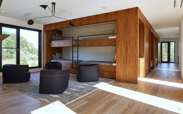 A bright contemporary multi-purpose room featuring built-in timber bunk beds and shelving, dark upholstered lounge chairs arranged on a large patterned rug, and extensive black-framed glazing. A timber-lined circulation zone and additional doors extend along the right side, while sunlight creates strong bands across the timber floor.