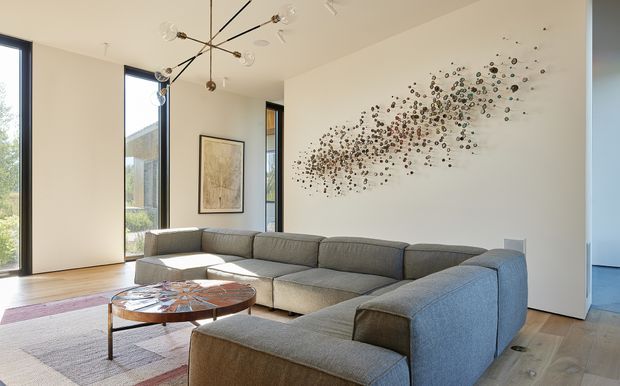 Bright contemporary living room with a large grey sectional sofa, low sculptural coffee table, pale timber flooring, tall narrow windows, and a white feature wall decorated with an abstract metallic installation. A branching modern chandelier and framed artwork add visual interest.