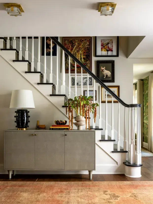 A refined residential hallway and stairway with a white-painted staircase, turned balusters, dark handrail and contrasting dark stair treads. A taupe sideboard with decorative objects sits beneath the stairs, while framed artwork, a black table lamp, indoor plants and a patterned rug add warmth and character. The adjoining room is partially visible through an opening on the right.