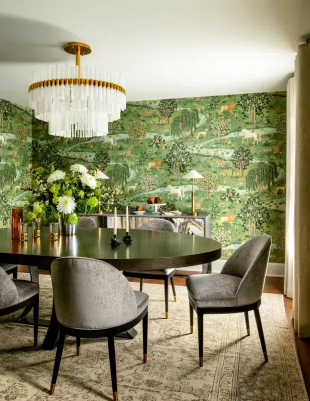 A refined dining room featuring a large round dark dining table surrounded by upholstered grey dining chairs, set over a patterned area rug. The room has a dramatic green botanical mural wallpaper, a brass-and-crystal tiered chandelier, a slim sideboard, decorative lamps, and a floral arrangement. Warm timber flooring and neutral curtains complete the richly layered interior.