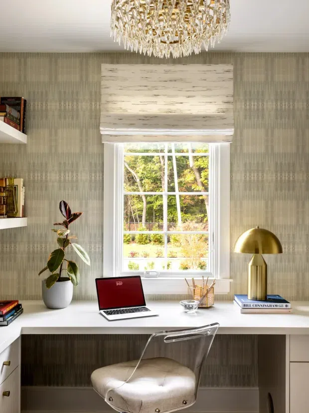 A refined home office arranged beneath a window, featuring a built-in white desk with drawers, a transparent acrylic chair, laptop, books, a potted plant, stationery, and a brass dome desk lamp. The room has patterned grey-green wallpaper, white trim, a textured Roman blind, and a dramatic crystal chandelier, creating an elegant, softly layered workspace.