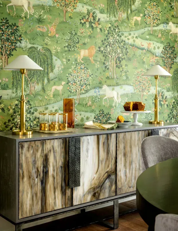 A richly styled interior vignette featuring a sculptural dark wood sideboard with dramatic pale and dark grain, topped with brass table lamps, glassware, plates, books, and a dessert stand. The wall behind is covered in a lush pastoral mural wallpaper depicting animals, trees, flowers, and a winding stream. A partial round dining table and upholstered chair are visible in the foreground. The composition has an eclectic, botanical, highly decorative character with warm metallic accents and strong material contrast.