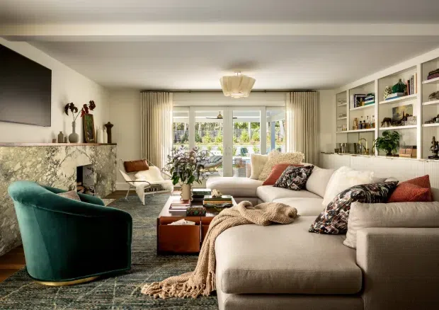 A warm, refined living room with a large cream sectional sofa, teal velvet accent chair, patterned rug, timber coffee table, marble fireplace surround, built-in open shelving, and expansive glazed doors leading to a garden. Neutral textiles are balanced by rust and teal accents, with layered curtains and a sculptural ceiling light.