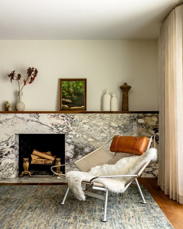 A refined living room centered on a dramatic natural-stone fireplace wall with a black firebox and timber mantel shelf. A mid-century style sling chair with a leather headrest and white sheepskin throw sits on a patterned area rug, while neutral curtains, warm timber flooring, ceramics, and artwork create a calm, sophisticated setting.