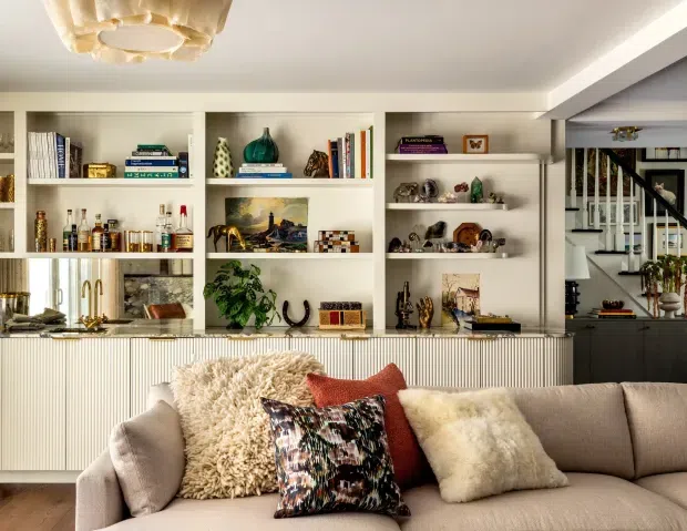Warm, refined living room with a large upholstered sofa in the foreground, layered textured and patterned cushions, and extensive built-in white display shelving filled with books, ceramics, artwork, and decorative objects. A ribbed white built-in cabinet runs beneath the shelving, while a staircase and hallway are visible at the right. The room has a softly lit, collected contemporary interior with an ornate ceiling light and neutral beige, white, and earthy accents.