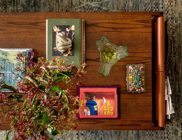 Overhead view of a richly styled wooden sideboard or console table, with visible wood grain, several art and design books, a sculptural glass bowl, a decorative mosaic-like object, a rolled leather-toned accessory, and a flowering branch. The table sits on a textured patterned rug, creating a warm, eclectic vignette.