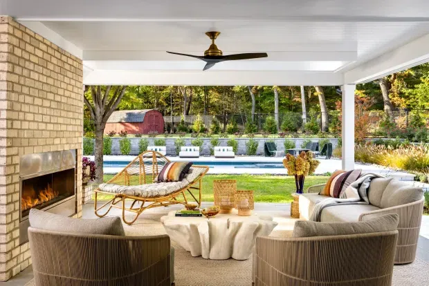 Bright, open-sided outdoor living room overlooking a landscaped garden and swimming pool. The space features a white paneled ceiling with a ceiling fan, a full-height cream brick fireplace, woven lounge chairs and sofa, a sculptural light-coloured coffee table, and a relaxed natural palette.