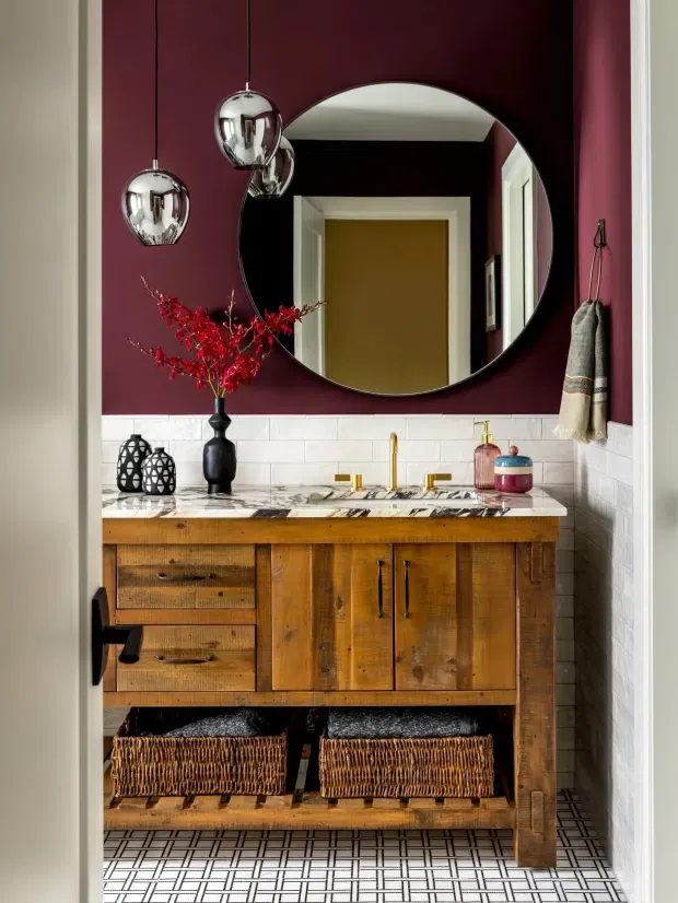 A richly styled bathroom vanity viewed through an open doorway. The room features a dark burgundy painted upper wall, white subway-tile wainscoting, a timber vanity with drawers and cupboard doors, a veined stone benchtop, brass-toned tapware, a large round mirror, chrome pendant lights, woven baskets, and patterned geometric floor tiles.