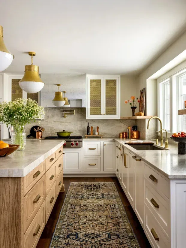 A bright, refined galley-style kitchen with a large central timber island, white shaker cabinetry, pale stone benchtops and backsplash, brass hardware and fixtures, and pendant lighting. A patterned runner adds warmth along the dark timber floor, while large windows provide abundant natural light.