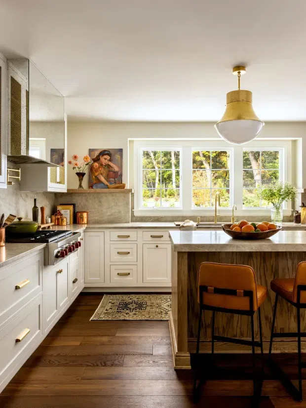 A warm, traditional kitchen with white shaker-style cabinetry, a marble-look backsplash and benchtop, dark timber flooring, and a timber-clad island with seating for two. A large brass pendant light hangs above the island, while generous windows overlook leafy greenery. Stainless steel appliances and decorative accessories complete the inviting interior.
