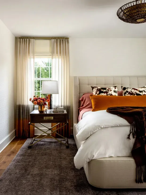Warm, refined bedroom interior featuring a fully upholstered, channel-tufted headboard, layered white and blush bedding, a rust-orange accent cushion, patterned pillows, dark throw, and a dark wood bedside table with crossed metal supports. Floor-to-ceiling curtains frame a window, while a woven pendant light and dark rug add texture. The room has white walls, timber flooring, and a soft, sophisticated palette.