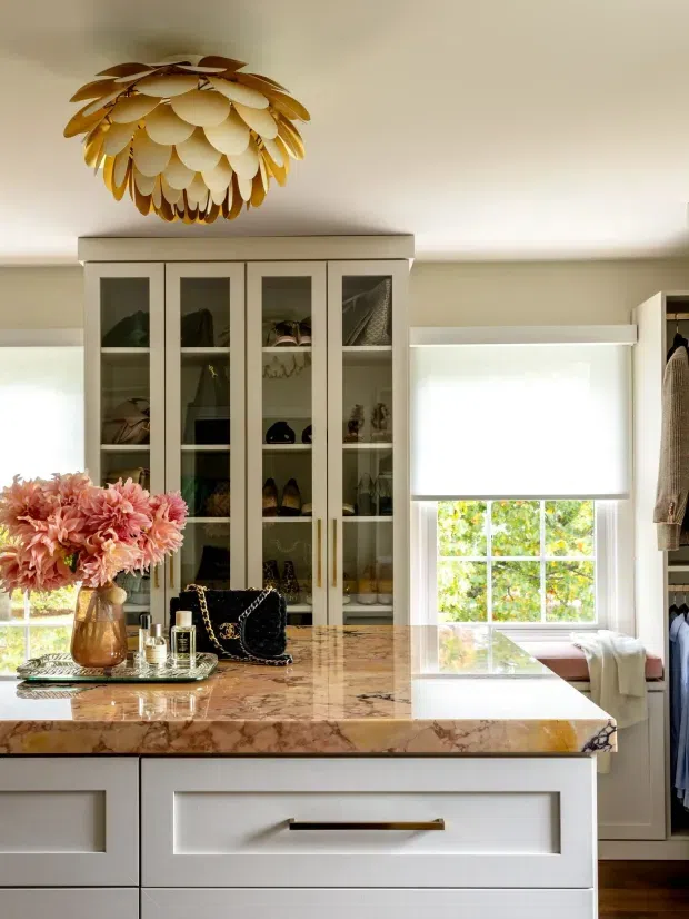 A refined walk-in wardrobe or dressing room with extensive cream built-in glass-front cabinetry, a large polished natural-stone island with drawers, clothing storage, and a decorative gold-toned pendant light. The space has bright windows with roller blinds, warm timber flooring, and elegant dressing accessories.