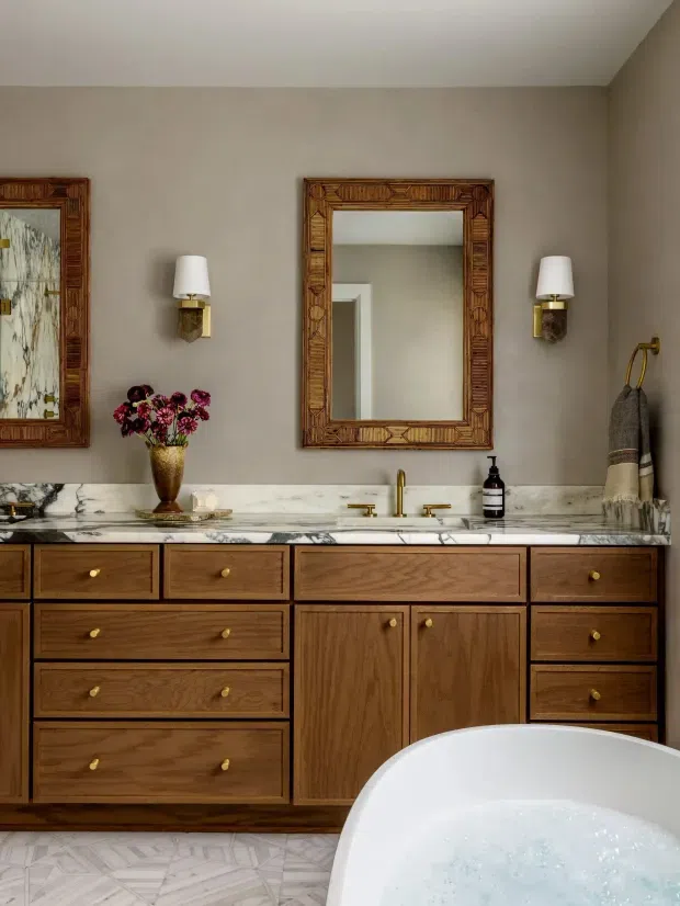 A refined bathroom interior featuring a large timber vanity with abundant drawers and brass knobs, a white marble countertop with integrated sinks and brass tapware, a freestanding white bathtub in the foreground, and warm taupe walls. Decorative carved timber-framed mirrors, wall sconces, folded textiles, and floral arrangements add a traditional, luxurious character. The floor is finished in a pale geometric stone or tile pattern.