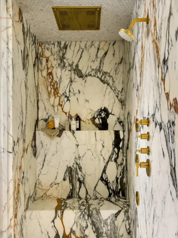 A compact, luxurious walk-in shower enclosed entirely in dramatic white marble with bold black, grey and warm ochre veining. The marble continues across the walls, ceiling, built-in shelf, and floor. Brushed brass/gold shower fittings, controls, and a ceiling-mounted shower vent create a refined high-contrast finish. Small toiletries sit on the recessed ledge.