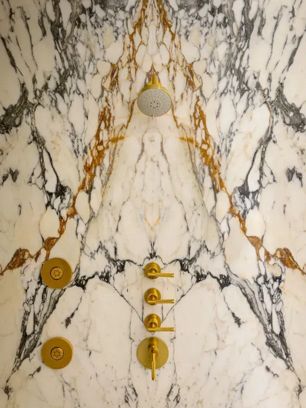 Close-up view of a luxurious shower wall clad entirely in dramatic white marble with bold black and warm gold veining. A central round showerhead and matching gold-toned controls and fixtures are symmetrically arranged across the stone surface.