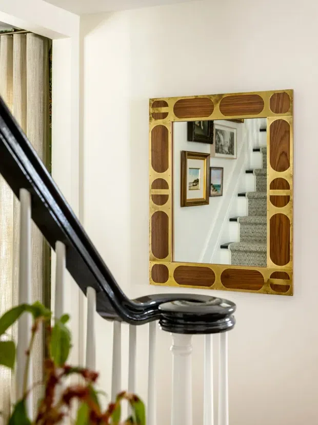 A close-up interior view of a stair landing or hallway featuring a decorative rectangular wall mirror with a geometric timber frame. The mirror reflects a carpeted staircase with white balustrades and framed artwork. A glossy black stair handrail and white spindles dominate the foreground, with light curtains and a small amount of greenery at the side.