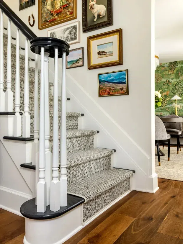 A traditional residential stairway with a curved black handrail and white turned balusters, patterned carpet runner, and dark timber flooring. The stair wall is decorated with a gallery of framed artworks, while an adjacent dining area with botanical wallpaper is visible through the opening.