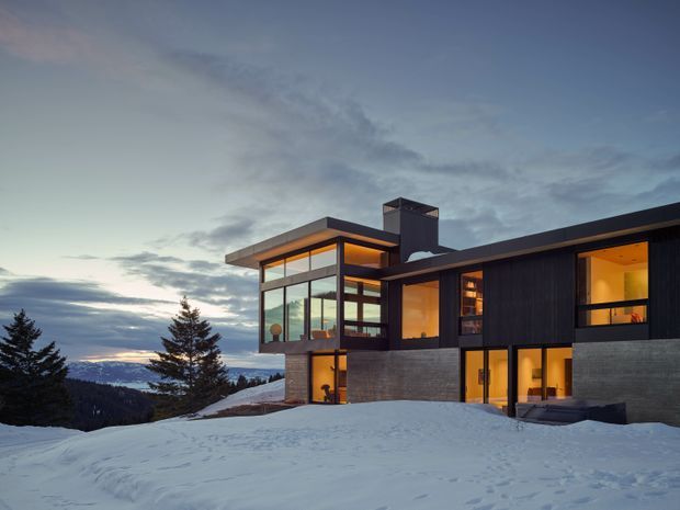A contemporary multi-level house set on a snowy mountain or alpine site at dusk. The dark horizontal facade and concrete lower walls contrast with extensive warm-lit glazing, including a prominent glazed corner volume, flat roof planes, and a tall chimney or vertical structural element. Snow-covered terrain and distant mountain views surround the building.