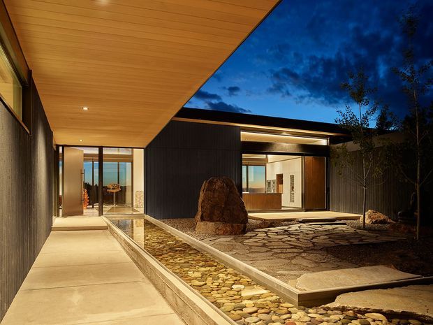 Twilight view of a contemporary single-level residence organized around a landscaped entry courtyard. A broad warm timber-lined soffit projects over a concrete walkway, with dark vertical wall cladding, extensive glazing, and a shallow pebble-lined water feature leading toward a large natural boulder. The courtyard includes irregular stone paving, integrated exterior lighting, and views into a bright interior living or kitchen area.