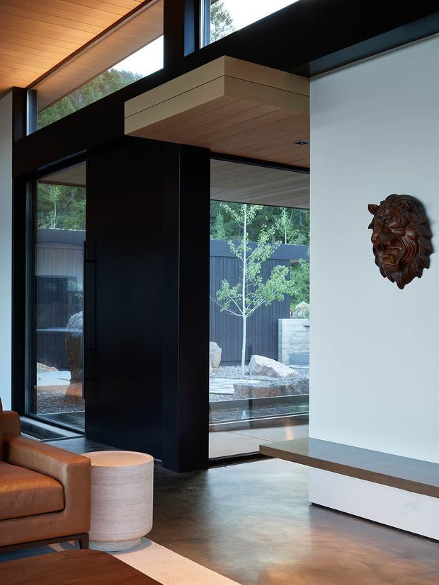 A refined contemporary living room with floor-to-ceiling glazing, dramatic black structural framing, warm timber-lined ceiling planes, polished concrete flooring, and a minimalist white built-in media wall. An enclosed landscaped courtyard is visible through the glazing. A caramel leather sofa and pale cylindrical side table add warmth, while a carved lion wall sculpture provides a decorative focal point.