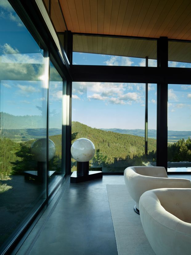 A refined modern living room with expansive floor-to-ceiling glazing overlooking a forested, mountainous landscape. Dark structural window mullions and framing contrast with a warm timber-paneled ceiling, polished concrete floor, and curved cream upholstered lounge chairs. A sculptural spherical light or artwork sits on a dark pedestal beside the glazing, while a pale area rug defines the seating zone. The space is bright, open, and strongly connected to the surrounding landscape.