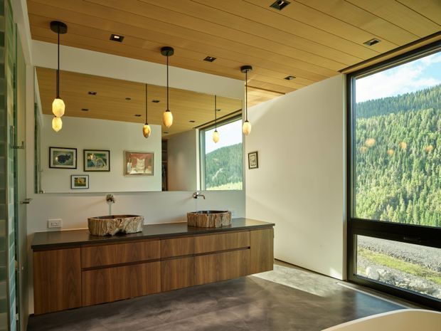 A contemporary bathroom vanity area with a long dark timber floating cabinet, two natural stone vessel basins, wall-mounted taps, and a large mirror. Warm timber ceiling boards, recessed downlights, and pendant lights create a refined atmosphere. Full-height black-framed glazing overlooks a forested mountain landscape, while polished concrete flooring and white walls keep the room bright and modern.