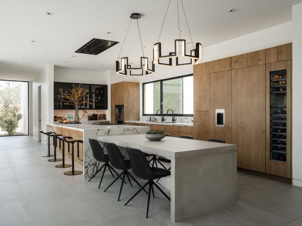 A spacious contemporary kitchen with a large central island featuring a light stone benchtop and concrete-look waterfall sides. The room has extensive warm timber cabinetry, integrated appliances including a tall wine fridge, a dark-framed window, pale large-format flooring, and a striking geometric black pendant light. Bar stools line a secondary peninsula, while black wire-frame dining chairs sit at the island.