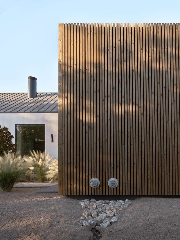 Exterior view of a contemporary building featuring a tall vertical timber screen or slatted wall in the foreground, with two circular outdoor fixtures near its base. Behind it is a low white-rendered structure with a dark metal pitched roof, chimney, dark-framed window, gravel landscaping, ornamental grasses, and a dry-stone drainage feature. The scene has clean modern lines and warm natural materials.