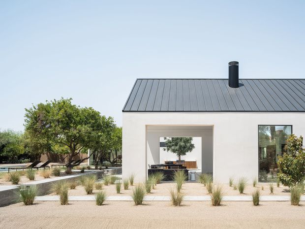 A minimalist single-storey building with smooth pale rendered walls and a dark standing-seam pitched roof, viewed from a landscaped exterior. A large square opening frames a visible outdoor dining or living area, while extensive gravel landscaping, concrete paths, ornamental grasses, mature trees, and a pool edge create a restrained contemporary courtyard setting.