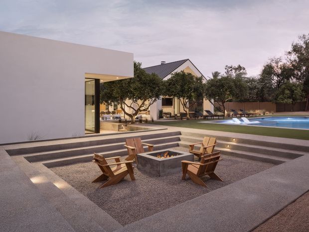 A contemporary residential outdoor entertaining area at dusk, centred on a square concrete fire pit surrounded by timber lounge chairs. Broad stepped concrete terraces lead toward a landscaped courtyard and a long swimming pool, with a minimalist white pavilion on the left and a pitched-roof house in the background. Integrated low-level lighting creates a warm, refined atmosphere.