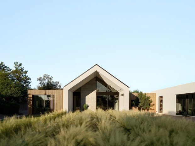 A contemporary single-storey residence viewed from landscaped grounds. The central gabled volume has a prominent triangular entry facade with deep roof overhangs, dark-framed glazing, and a stone or concrete-toned feature wall. Low flat-roofed wings extend to either side, including a timber-clad glazed section. Ornamental grasses and mature planting dominate the foreground.