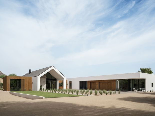 Wide exterior view of a low, contemporary residential complex arranged around a landscaped courtyard. The main gabled volume has white rendered walls, dark metal roofing, extensive black-framed glazing, and vertical timber cladding. A long flat-roofed wing extends to the right with large glazed openings and a timber-clad recessed facade. Sparse ornamental planting, paved ground, and a broad open forecourt create a clean, modern composition.