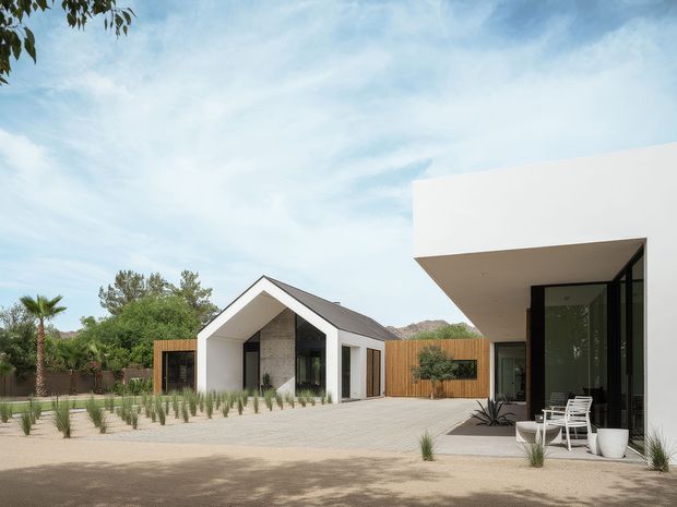 Exterior view of a contemporary residential property with a small white gabled pavilion and a larger flat-roofed volume connected by landscaped paving. The design features crisp white rendered walls, dark-framed glazing, timber screening, sparse drought-tolerant planting, and a sheltered outdoor seating area. A low mountain ridge and mature vegetation form the background.