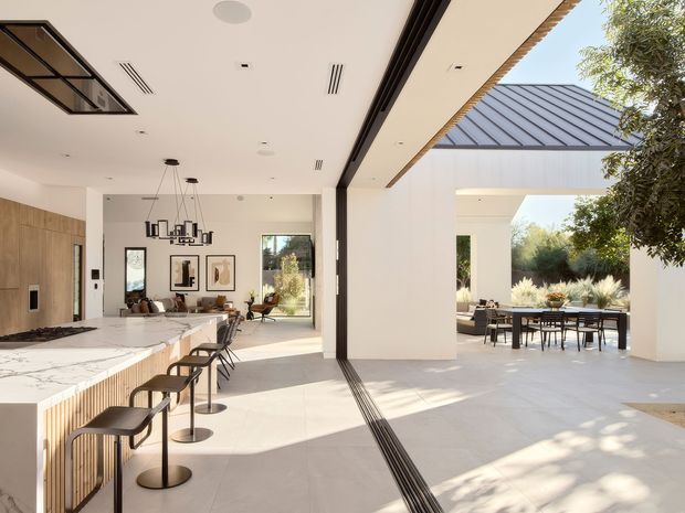 Bright, open-plan contemporary interior connecting a kitchen, living area, and dining terrace through wide sliding openings. The space features a large marble-look island with timber slatted cabinetry, dark-framed glazing and doors, polished light flooring, a linear black kitchen hood, and a covered outdoor dining area beyond. Abundant daylight and landscaping create a refined indoor-outdoor connection.
