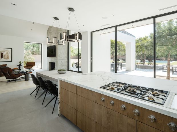Bright, open-plan contemporary kitchen with a large wood-fronted island, white veined stone benchtop, integrated gas cooktop, and black bar stools. The kitchen opens directly to a spacious living area and a covered outdoor patio through expansive black-framed glazing. A concrete feature wall with fireplace and sculptural pendant lighting adds architectural interest.