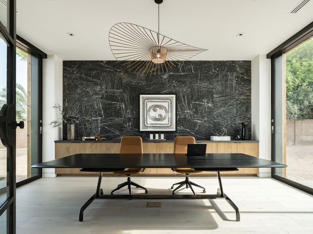 A refined contemporary dining or meeting space with a long black table and brown leather office-style chairs, positioned in front of a full-width dark textured stone feature wall. A low timber sideboard runs along the wall beneath framed artwork and decorative objects. Floor-to-ceiling black-framed glazing on both sides provides abundant natural light, while a sculptural linear pendant light hangs above the table. The room has pale large-format flooring and a restrained palette of black, timber, tan, and off-white.