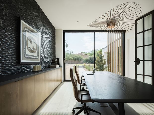 A refined contemporary dining and meeting space with a long dark timber table, office-style chairs, a light timber sideboard, textured black feature wall, framed artwork, and a sculptural oversized pendant light. Full-height glazing opens the room to a landscaped outdoor area, while a black-framed glazed door adds a strong architectural detail.